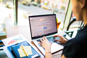 Person working on a laptop at a desk with charts and notes in a bright, windowed space.