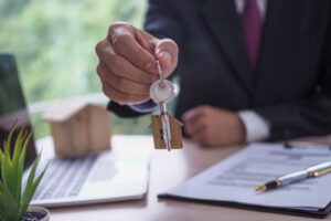 A person in a suit is holding out a key with a small house-shaped keychain, suggesting the handover of a property or real estate transaction. In the background, there is a contract, a pen, and a small house model on a desk.