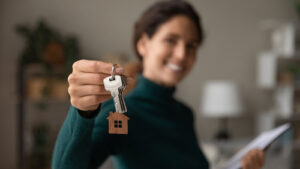Smiling woman holding up house keys with a wooden house-shaped keychain.