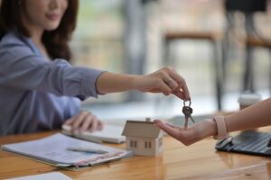  A person handing over house keys to another person across a desk with real estate documents and a house model, symbolizing a property transaction.