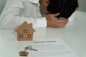 A person with their head in their hands, sitting behind an eviction notice, a house model, and a key, symbolizing housing distress.
