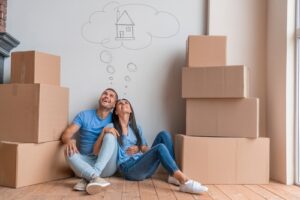  A happy couple sitting on the floor surrounded by moving boxes, looking up at a thought bubble with a house drawing inside.
