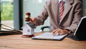 A person in a suit holding a gavel next to a house model, reviewing documents, symbolizing legal proceedings related to real estate or property.
