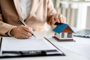 A person signing a document with one hand while resting the other on a small house model placed on the desk.