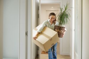 A woman carrying a moving box and a potted plant while talking on the phone, smiling as she walks through a hallway.