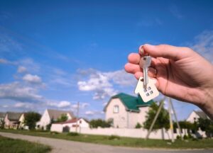 A hand holding a house-shaped keychain with keys, with a neighborhood of houses in the background.