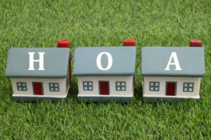 Three model houses on grass, each labeled with a letter to spell "HOA," representing a homeowners association.